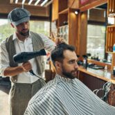 Een barbier met een klassieke outfit en een platte pet droogt het haar van een klant met een föhn in een traditioneel ingerichte barbershop. De klant draagt een gestreepte kapperscape en kijkt geconcentreerd vooruit.