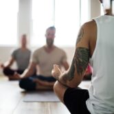 Een groep mannen volgt een Yin Yoga-les, met vooraan een deelnemer met tatoeage die in lotushouding zit. De foto laat zien hoe Yin Yoga rust en concentratie brengt zonder zweverig te zijn.