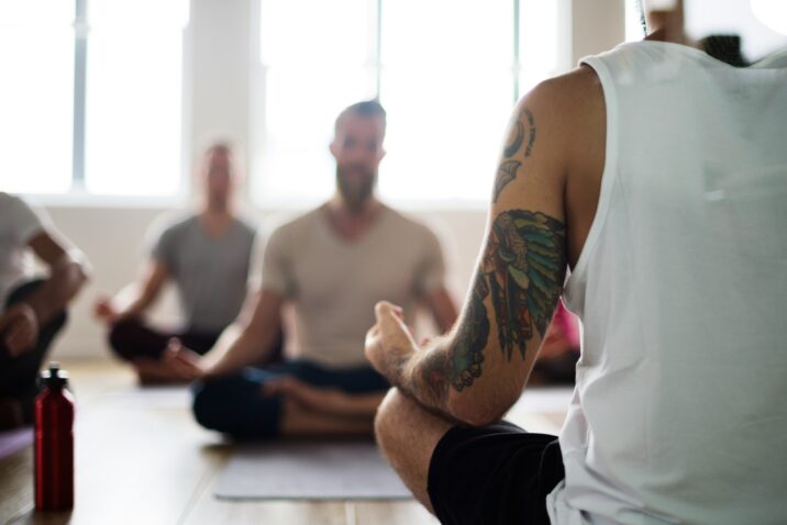 Een groep mannen volgt een Yin Yoga-les, met vooraan een deelnemer met tatoeage die in lotushouding zit. De foto laat zien hoe Yin Yoga rust en concentratie brengt zonder zweverig te zijn.